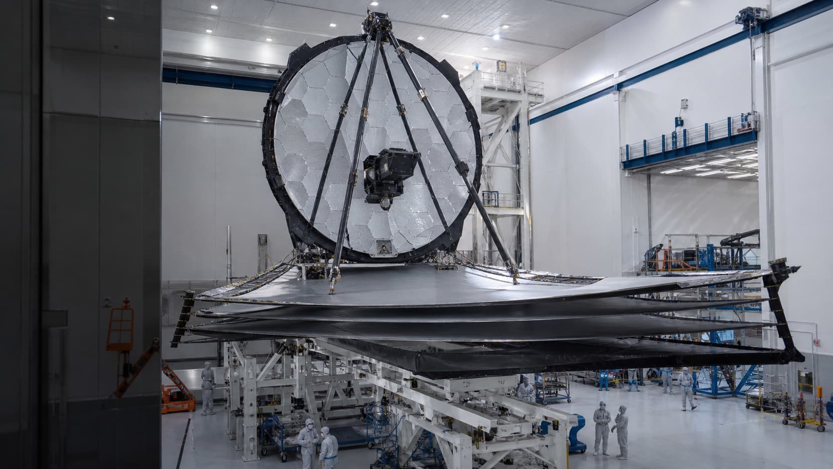A photorealistic space telescope being prepared inside a vast clean room under cool work lights